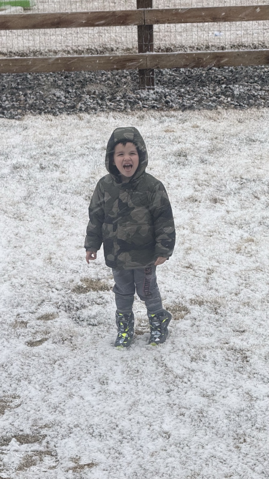 Luke in a camo hooded coat and snow boots, mouth wide open, laughing in falling snow