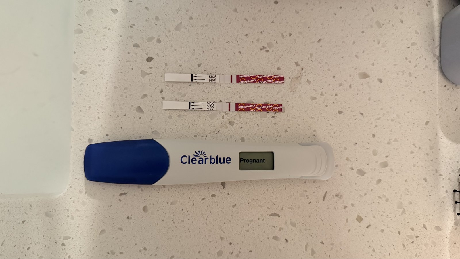 Two Pregmate pregnancy test strips with faint second lines, next to a Clearblue digital test reading Pregnant, laid out on a kitchen counter